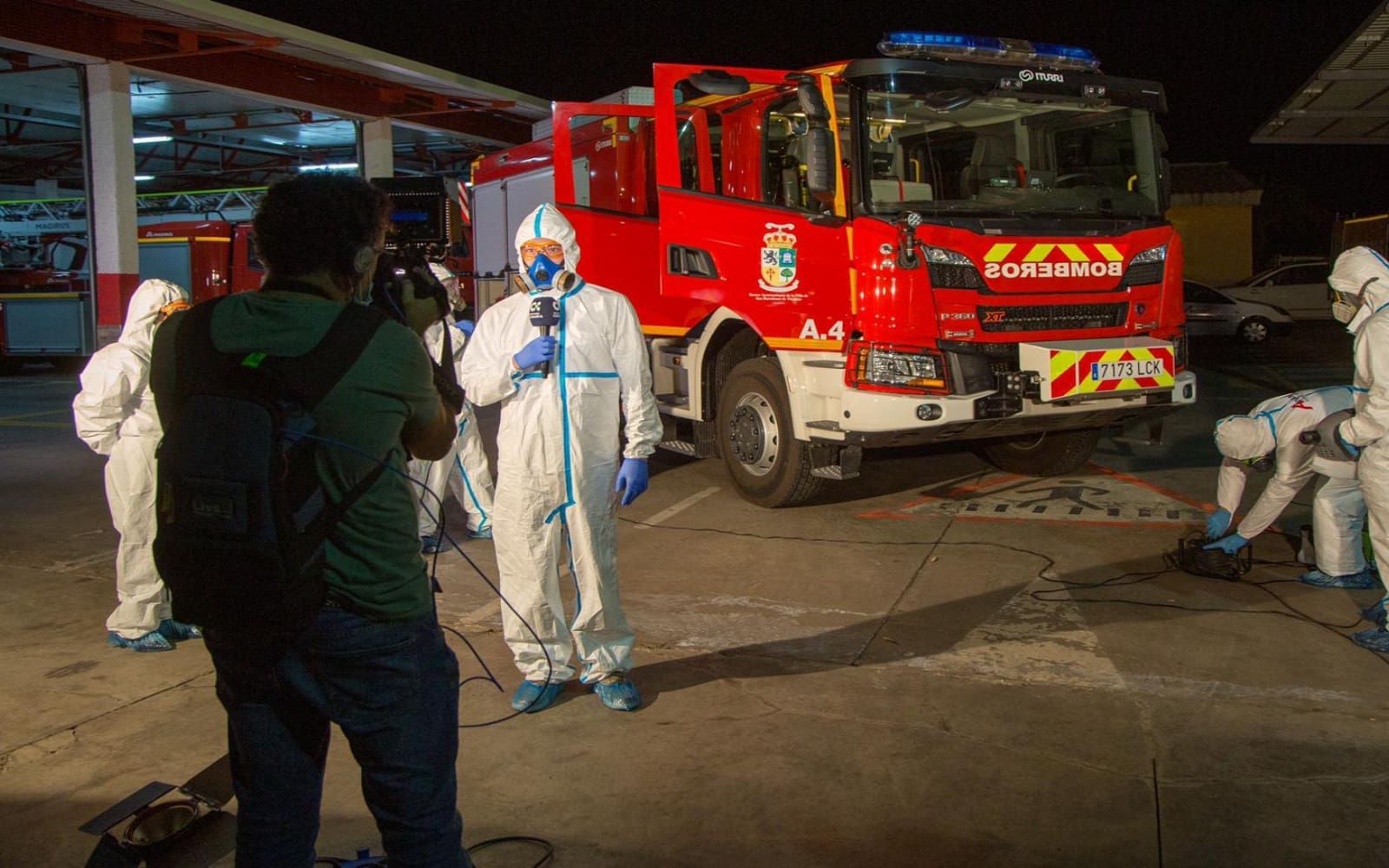 Personal de anviana con un traje epi delante de un camión de bomberos mientras lo graba la televisión.
